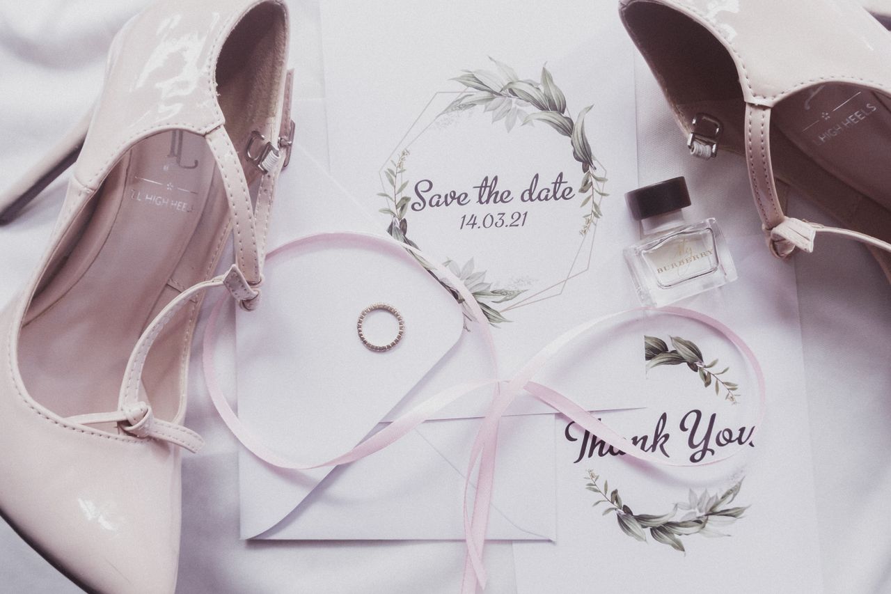 Une élégante composition à plat d’éléments essentiels pour un mariage, avec des talons, du parfum et des invitations.
